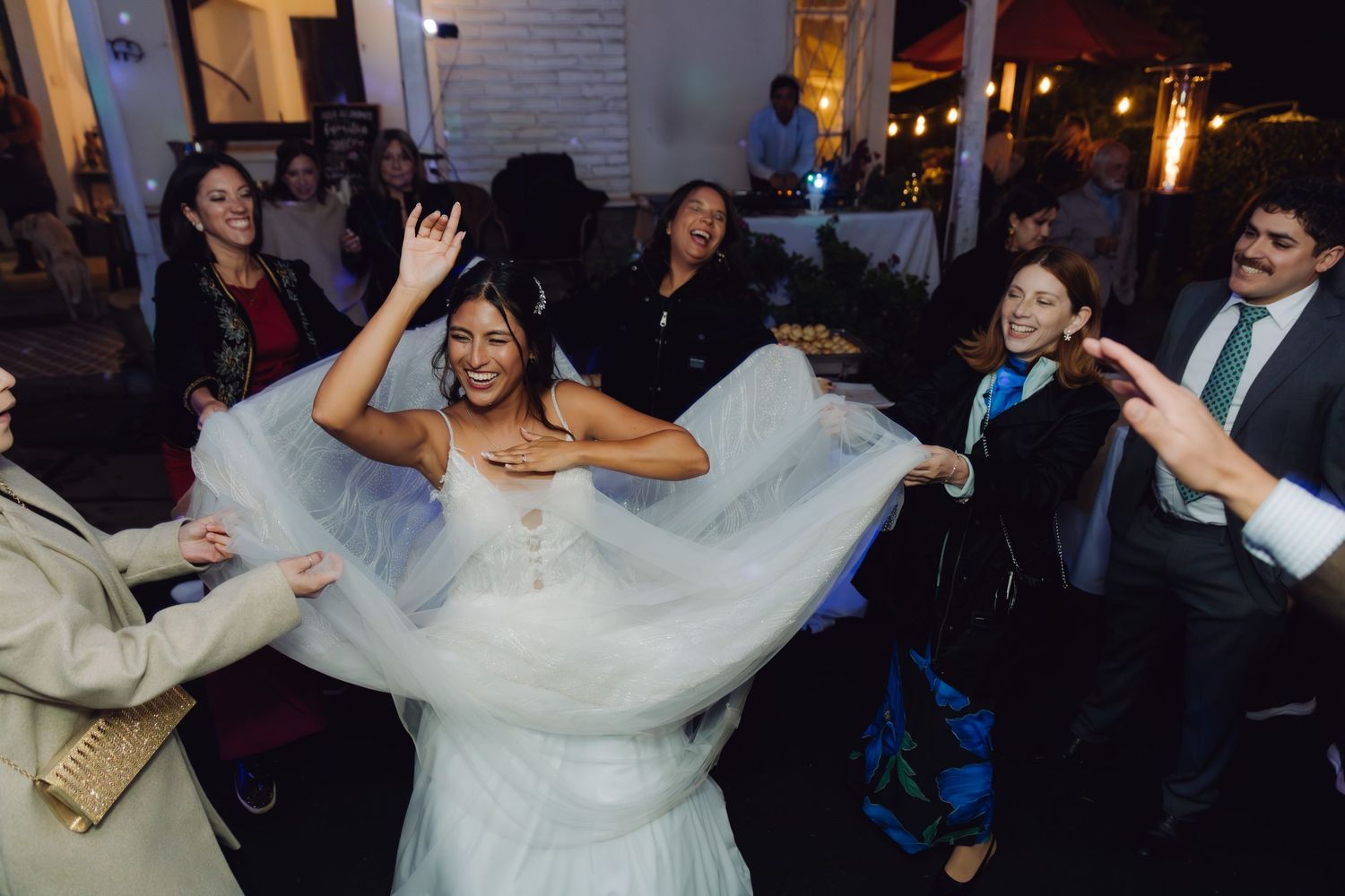 Bride in white tulle gown dances joyfully as guests spread her veil at wedding reception.