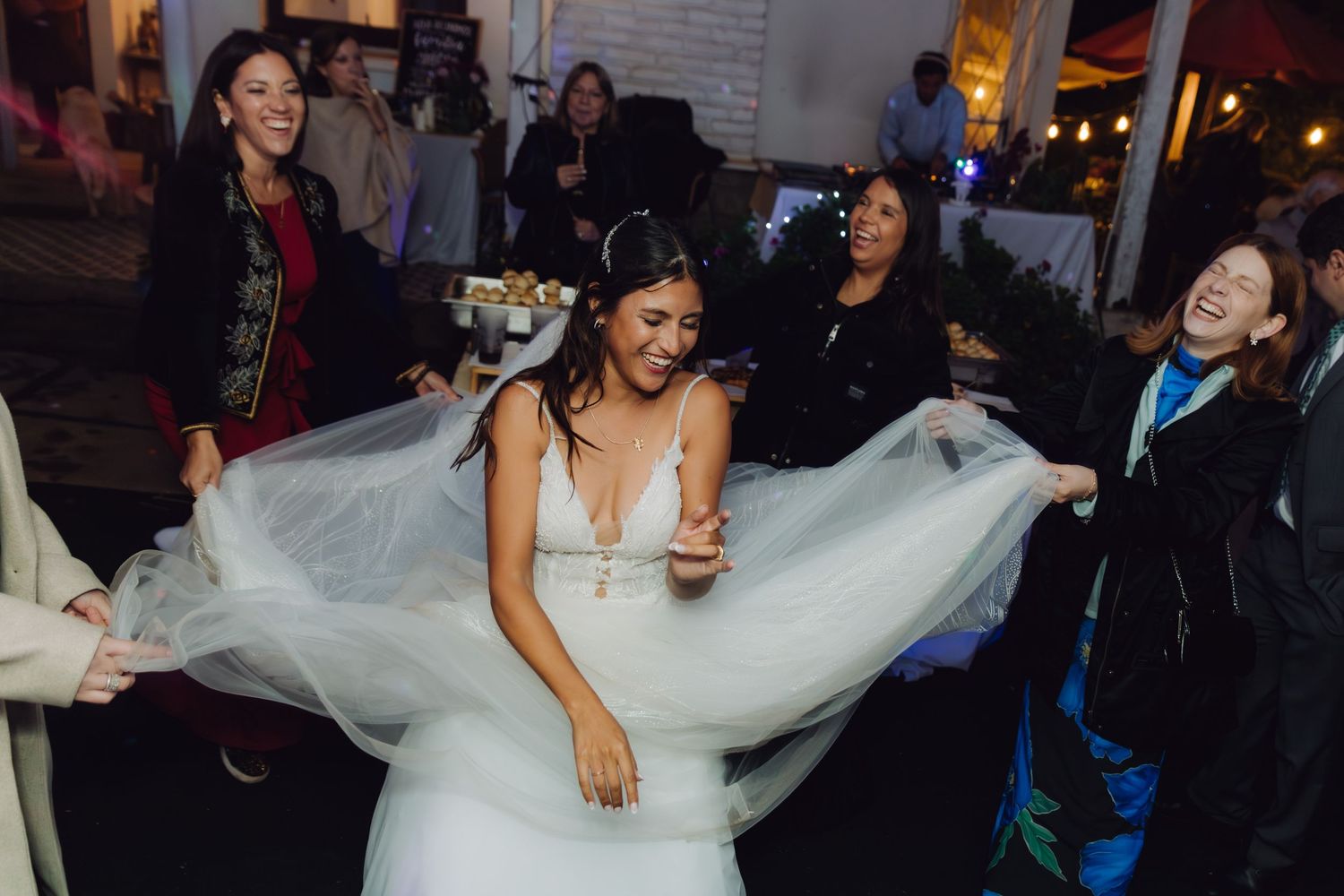 Bride in white gown laughs while guests hold her veil spread wide at wedding reception.