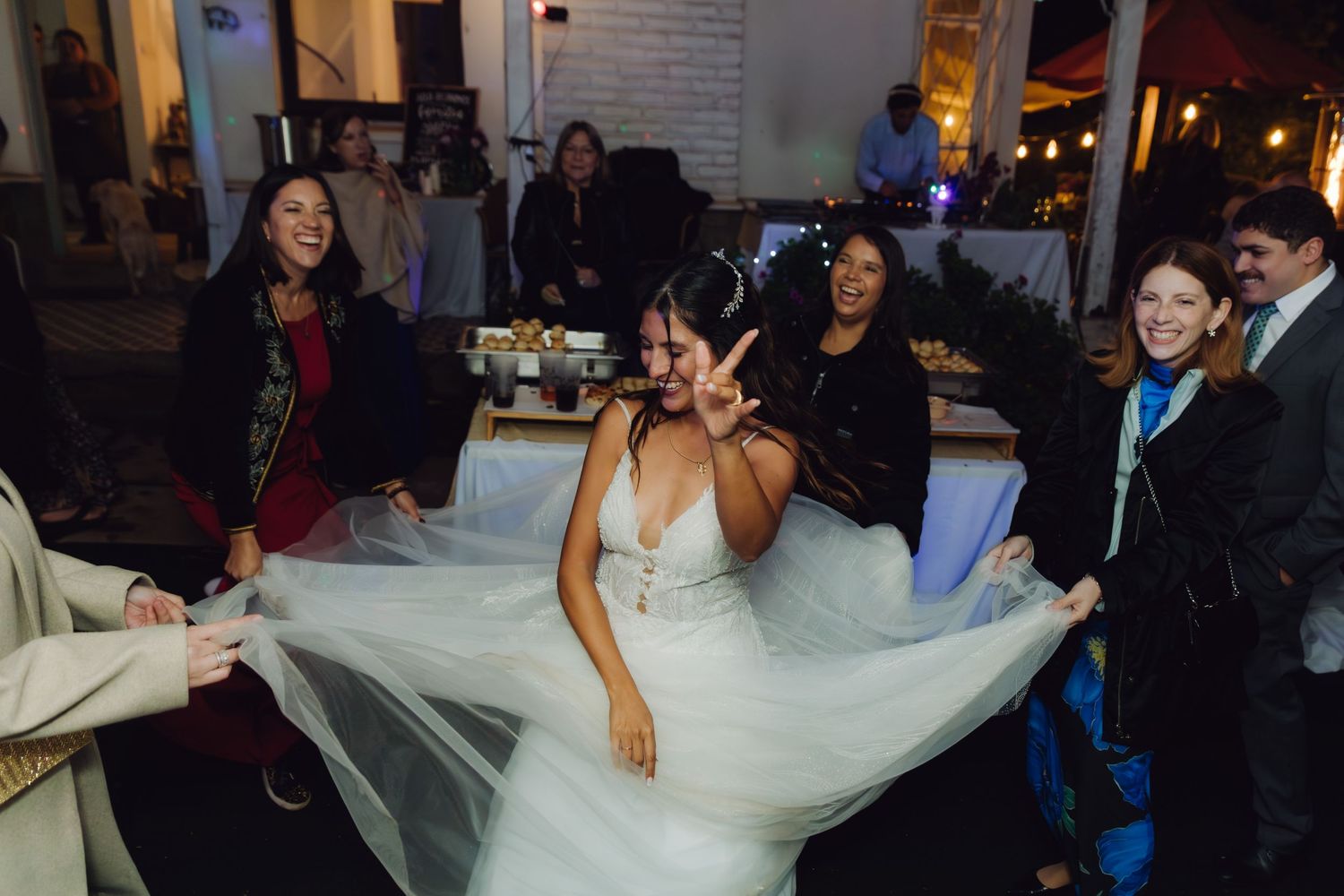 Bride in white gown flashes peace sign as guests hold her veil at lively wedding reception.