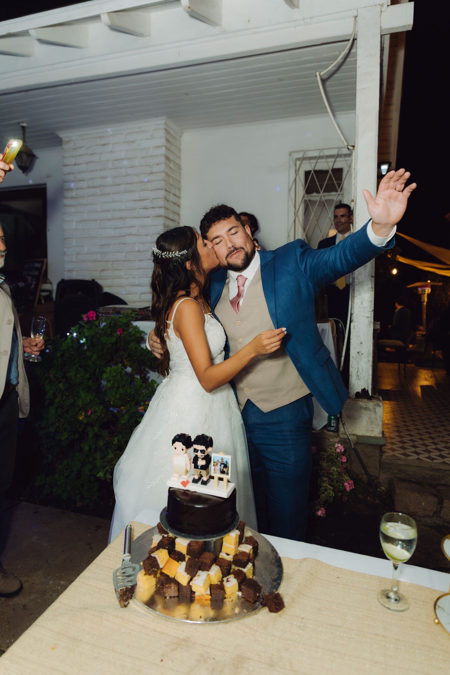 Bride kisses groom on cheek while cutting wedding cake at outdoor reception.