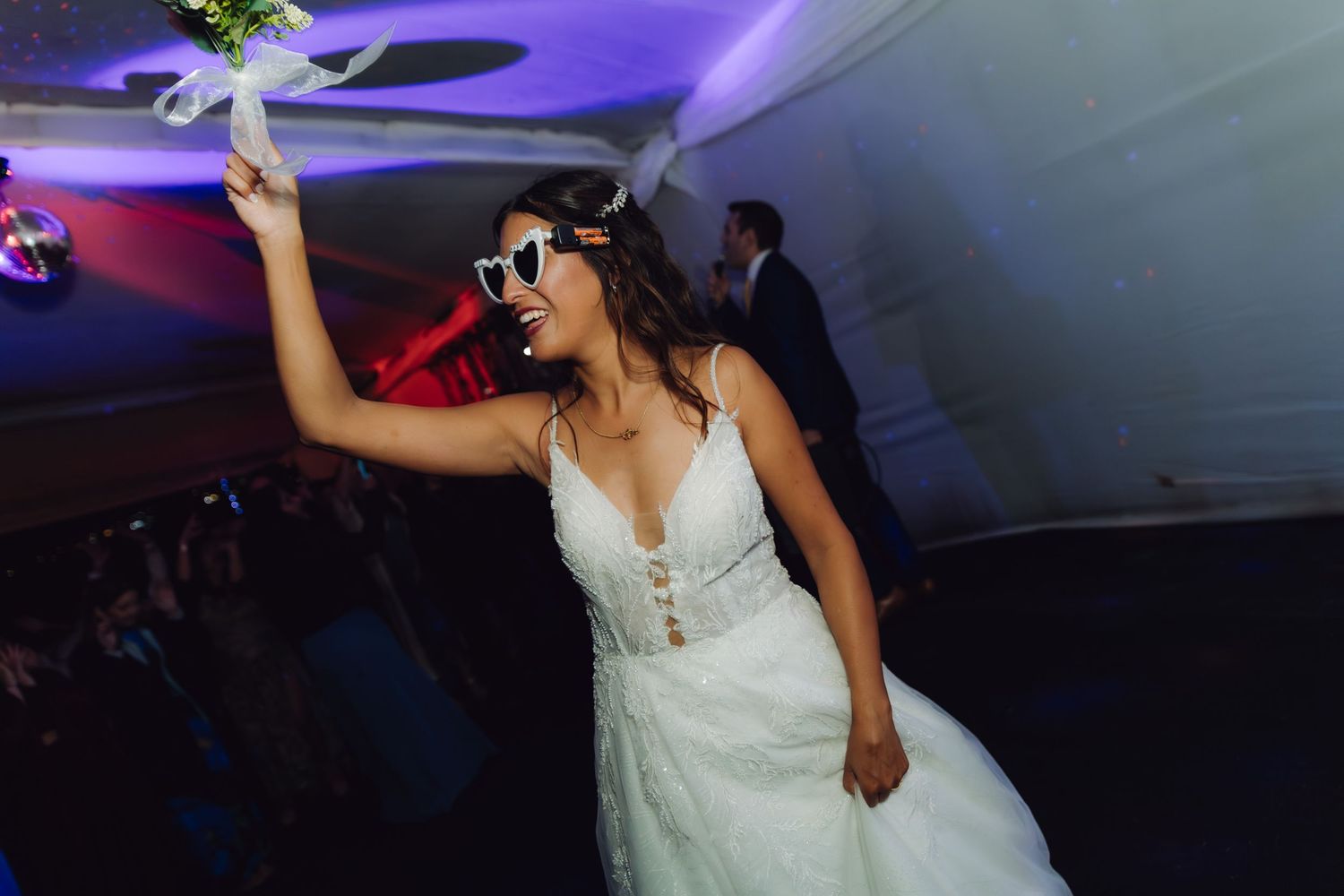 Bride in white gown tosses bouquet wearing heart sunglasses at wedding reception.