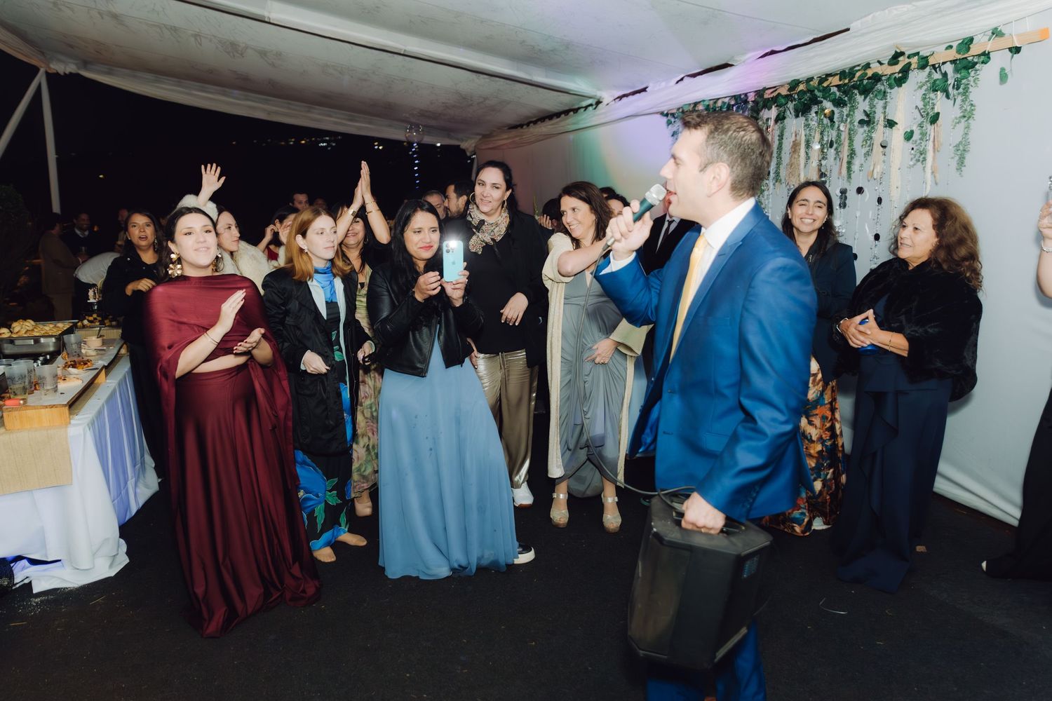 Groom in blue suit entertains cheering wedding guests on dance floor.