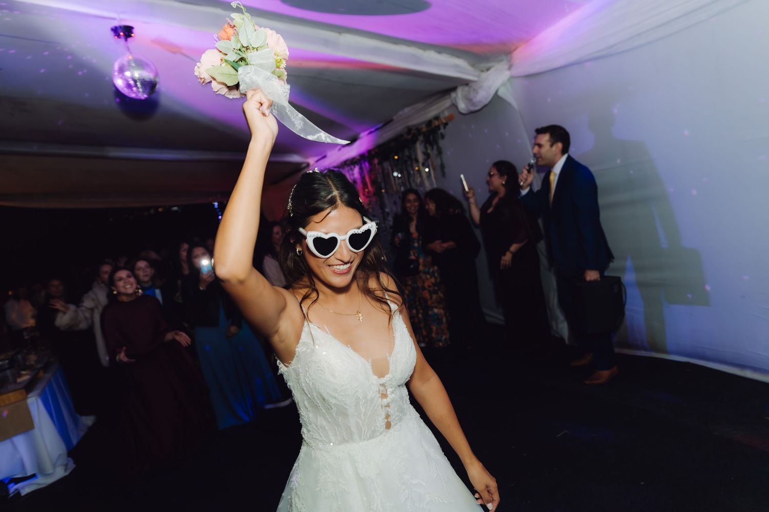 Bride in white gown raises bouquet wearing heart-shaped sunglasses on dance floor.