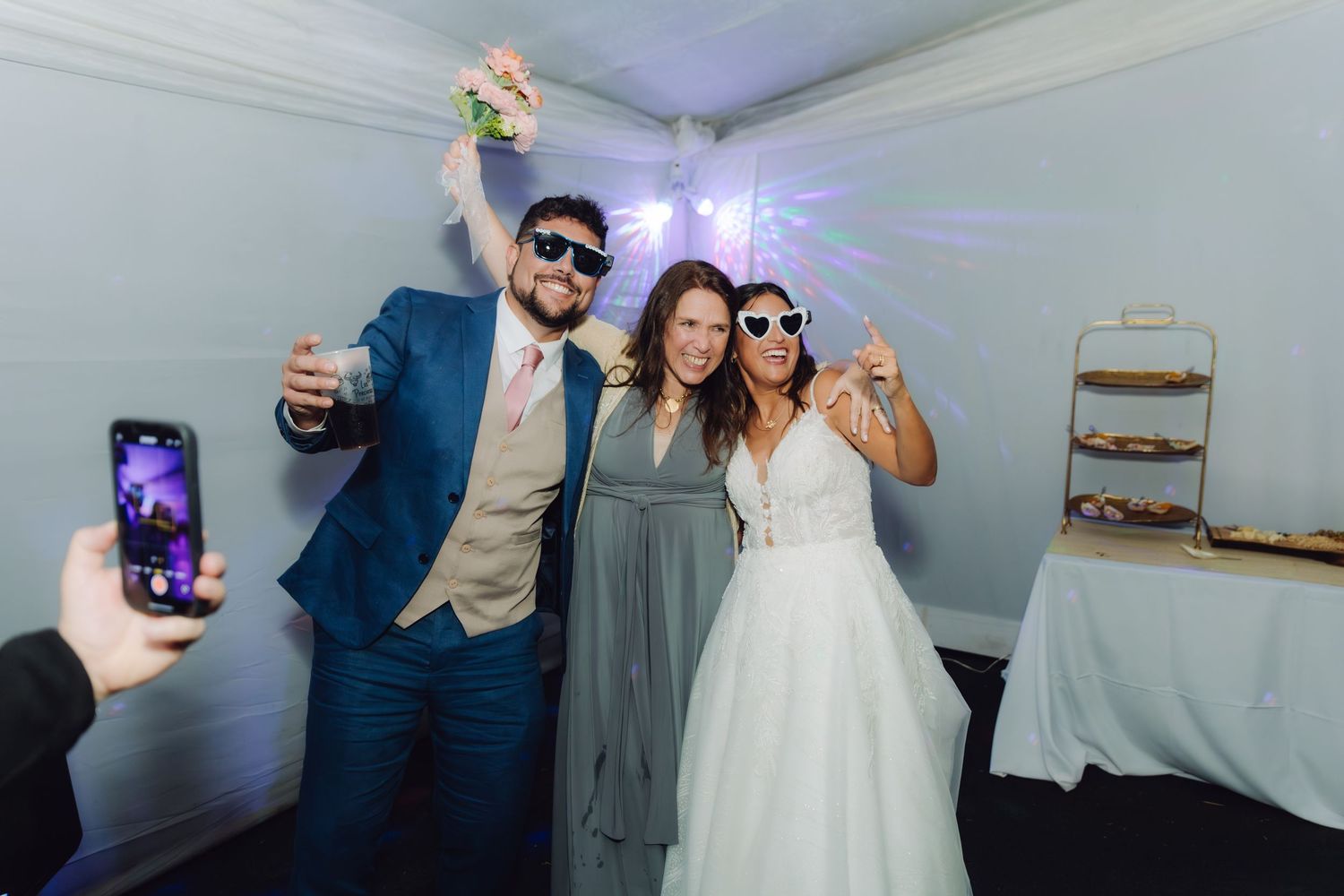 Bride, groom, and guest posing with sunglasses for a selfie at a wedding reception.