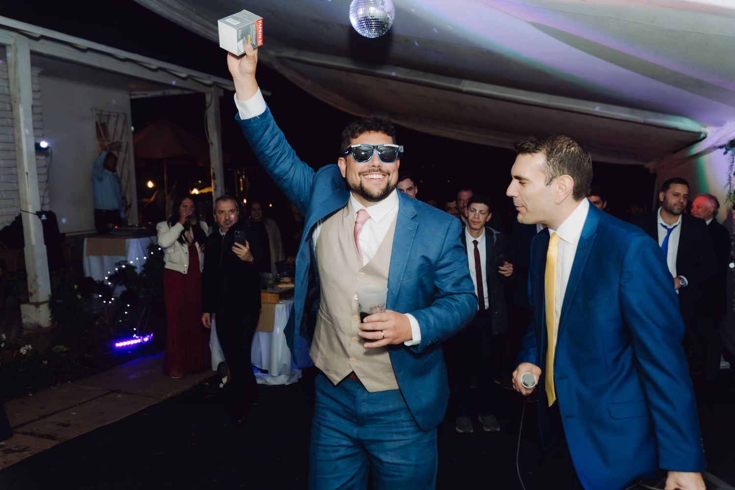 Groom in blue suit wearing sunglasses dancing energetically at his wedding reception.