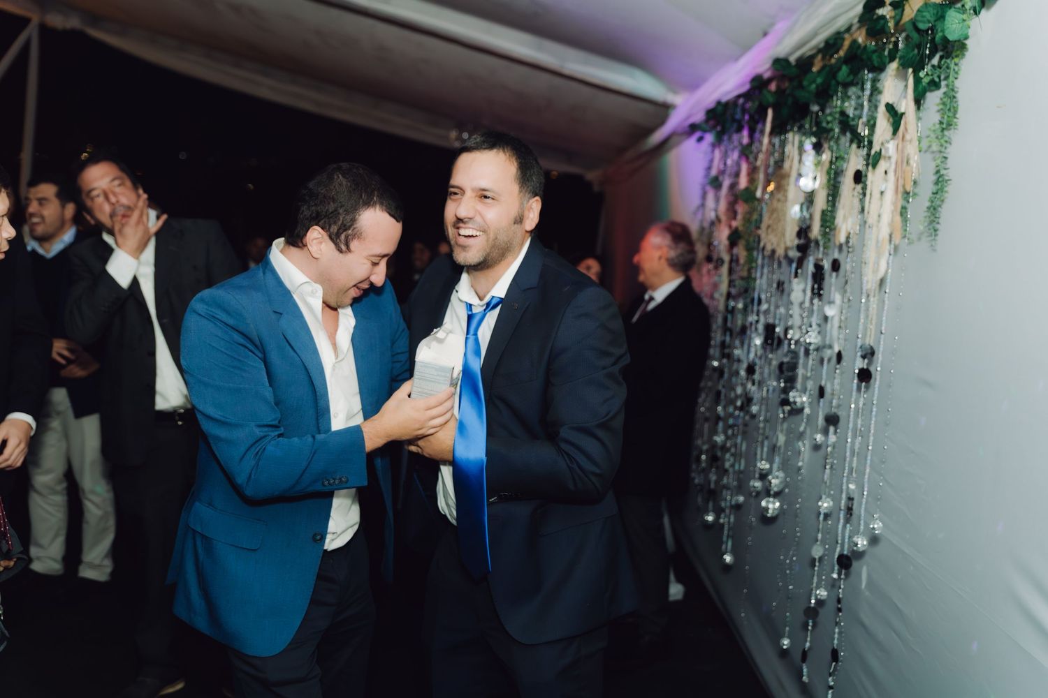 Two men in suits smiling and chatting at a decorated wedding reception venue.
