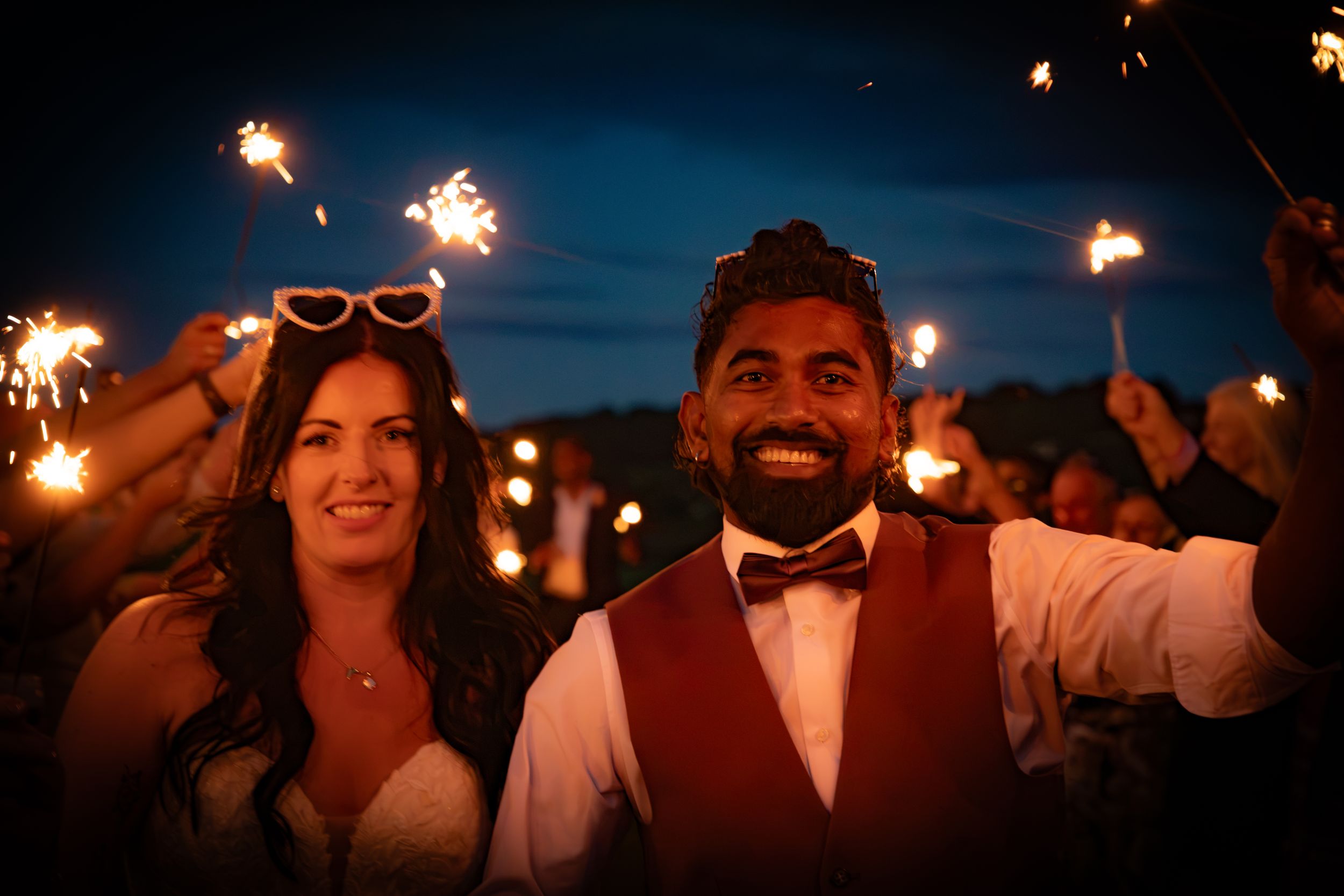 Bride and groom celebrating with sparklers at night, bride wearing heart-shaped sunglasses, groom in bow tie.