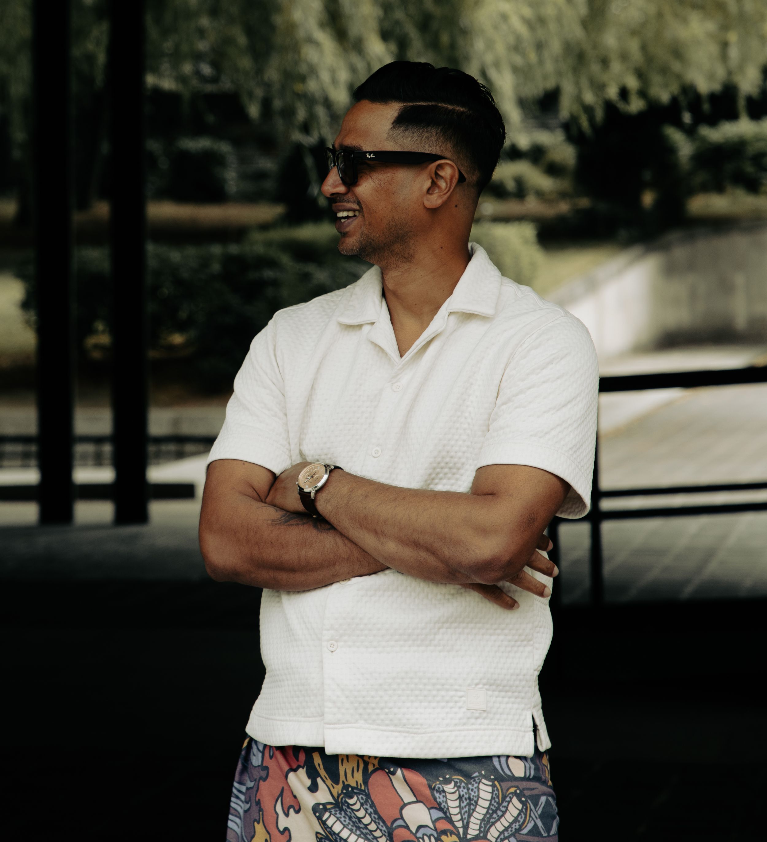 Stylish man in white knit shirt and patterned shorts posing with arms crossed wearing sunglasses outdoors.