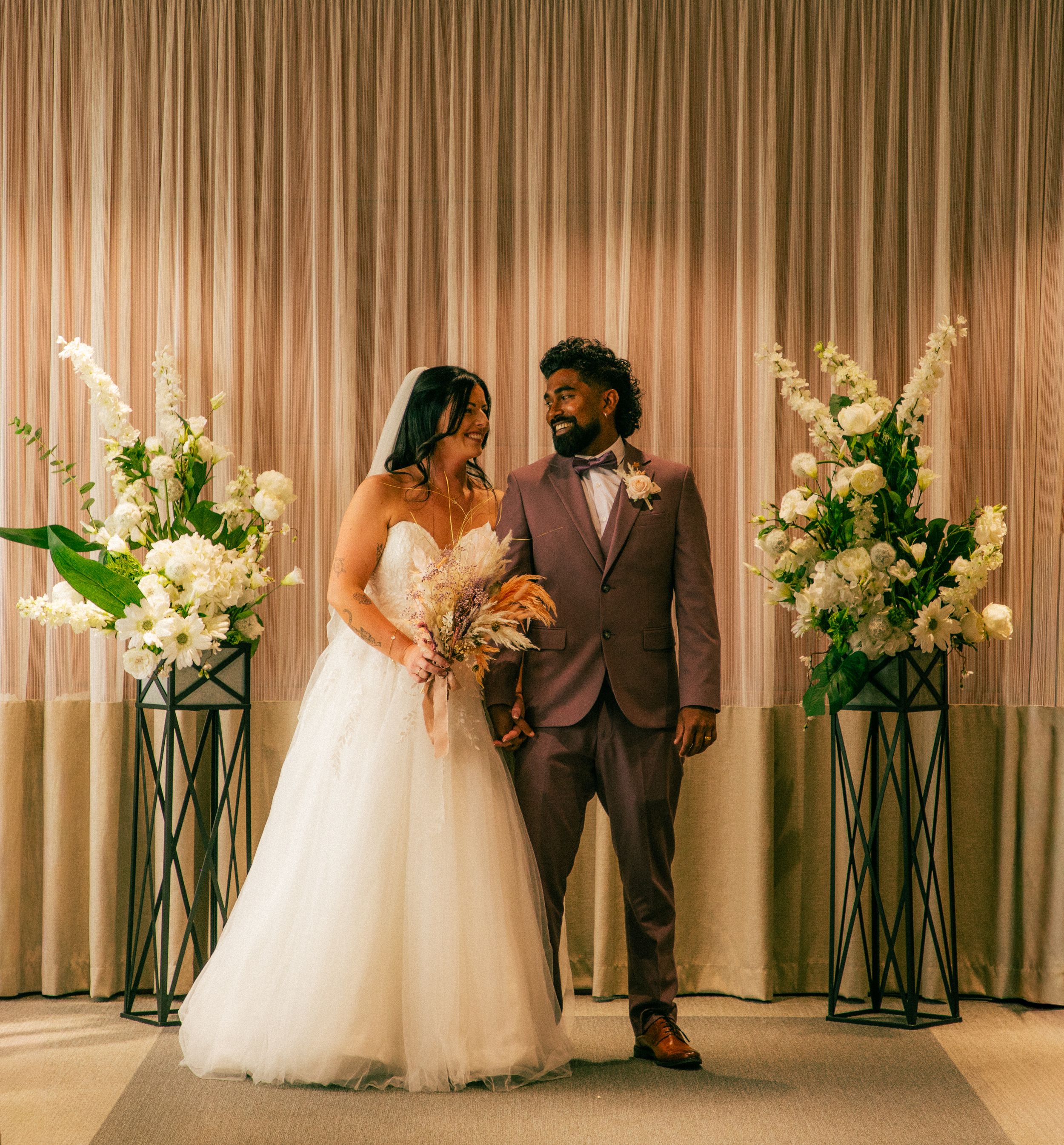 Bride in white tulle gown and groom in mauve suit standing together at a floral wedding ceremony altar.