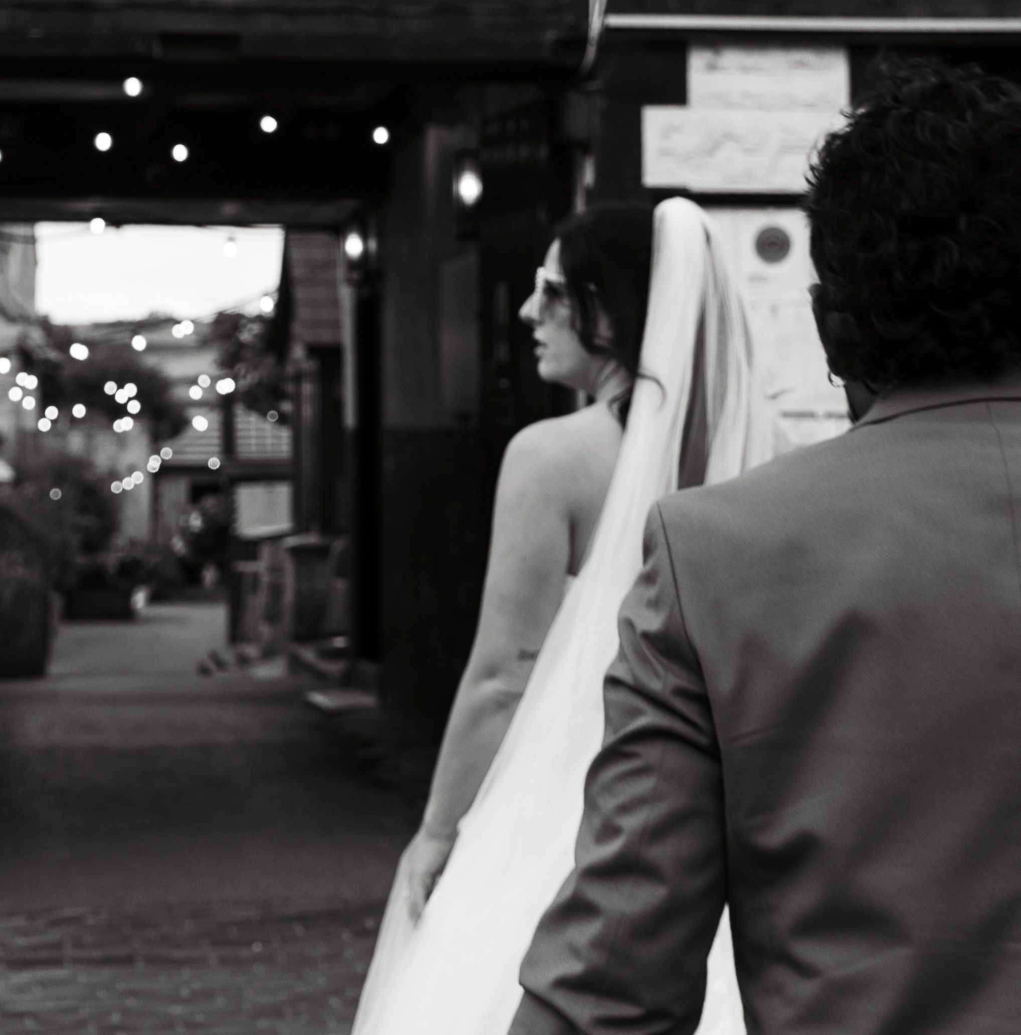 Black and white photo of a bride in a white gown and veil walking with a groom in a suit along a decorated outdoor venue.