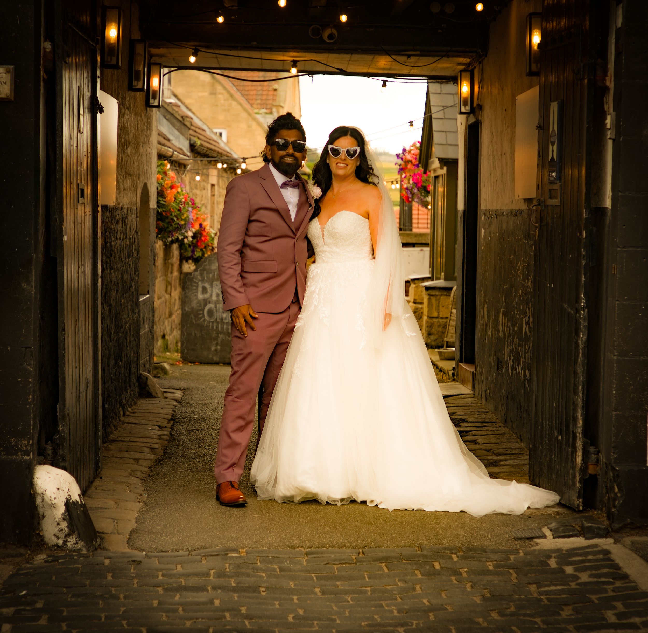 A bride and groom in stylish wedding attire pose in a charming cobblestone alley with festive string lights.
