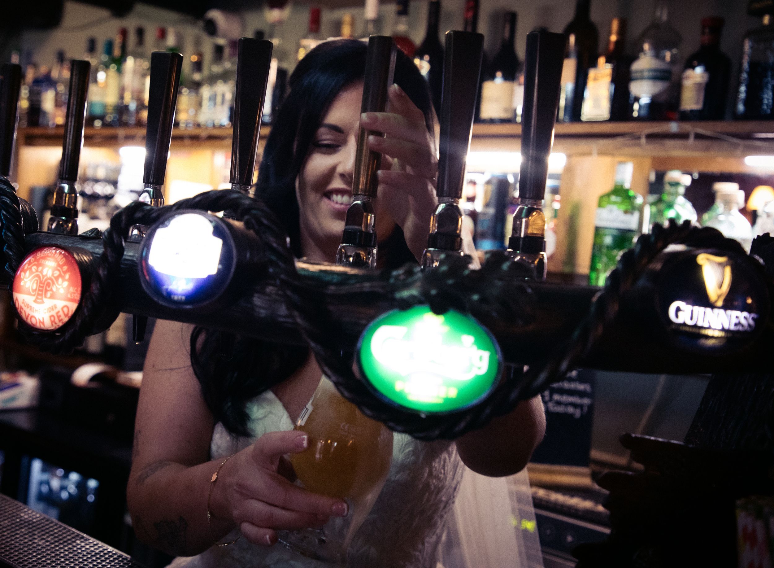 A bride in a white wedding dress smiles while pouring a pint of Guinness at a pub bar.