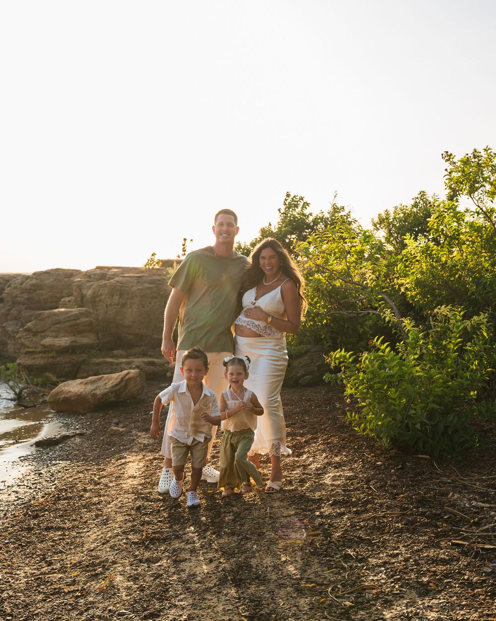 A happy family of four walks outdoors near rocks and greenery in warm golden sunlight, with a pregnant mother in white.