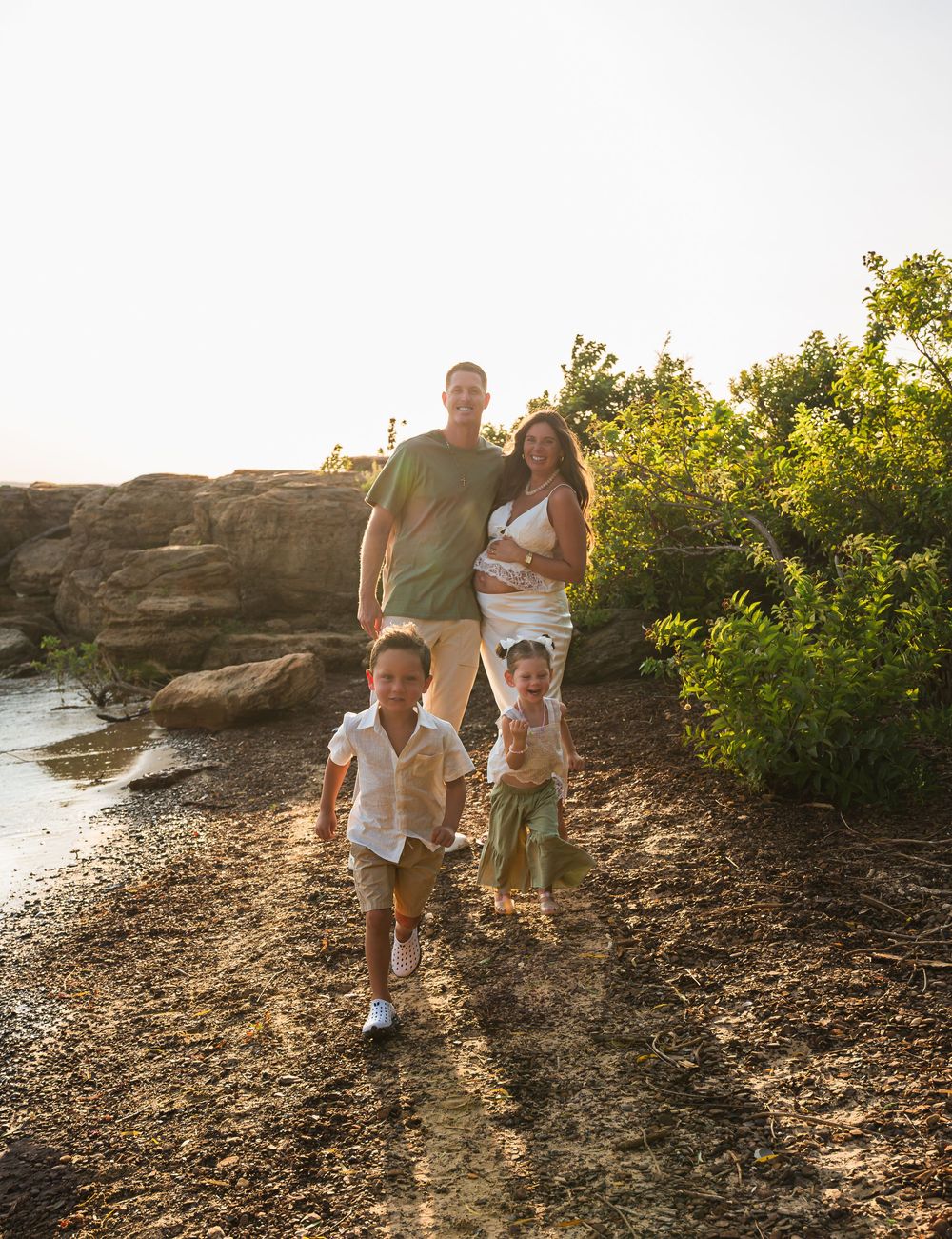 Family of four enjoying a sunny coastal walk near rocky shoreline and lush greenery during golden hour.