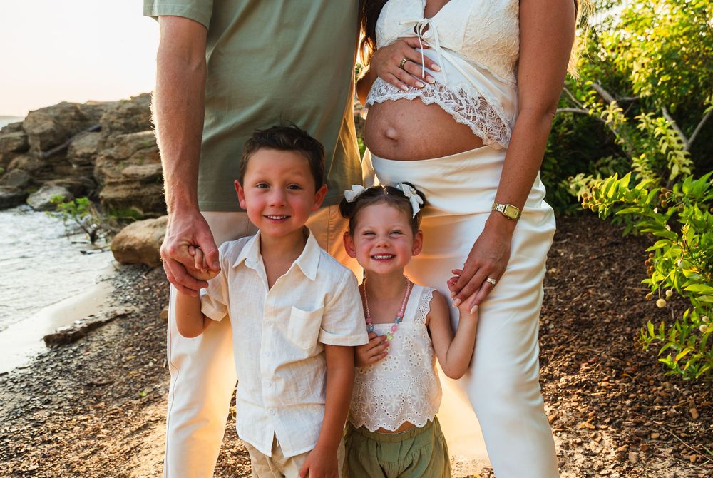 Pregnant woman in white lace top poses outdoors with two smiling children near rocky waterfront at golden hour.