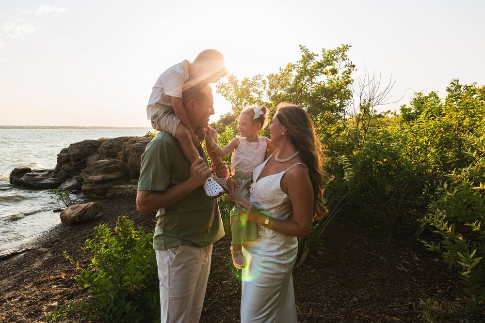 Family with two young children enjoying a golden sunset by the rocky shoreline surrounded by lush greenery.