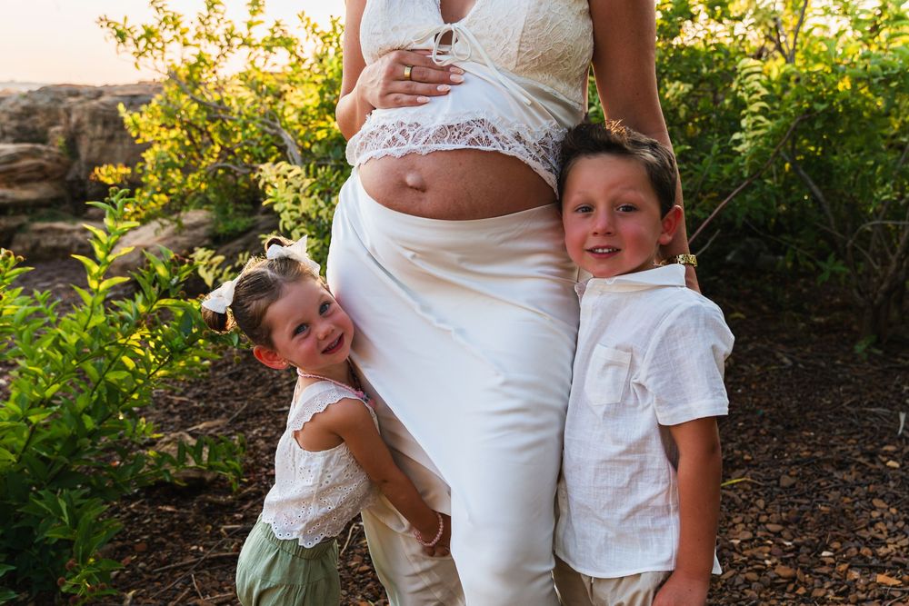 Pregnant woman in white outfit poses outdoors with two young children hugging her belly during a maternity photo session.