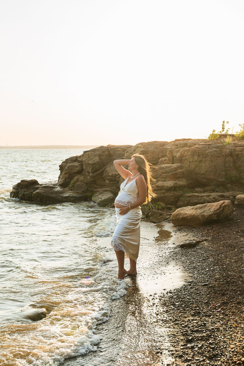 Pregnant woman in white dress standing barefoot on rocky shoreline at sunset, cradling her belly by the ocean.