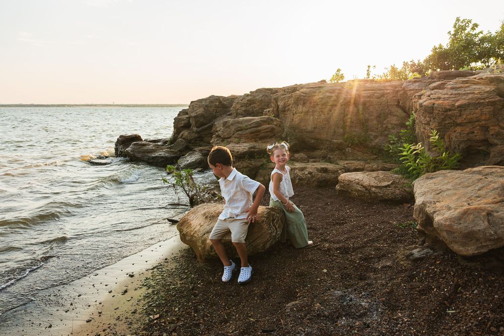 Two children sitting on rocky shoreline at golden hour sunset, wearing white outfits near calm water.