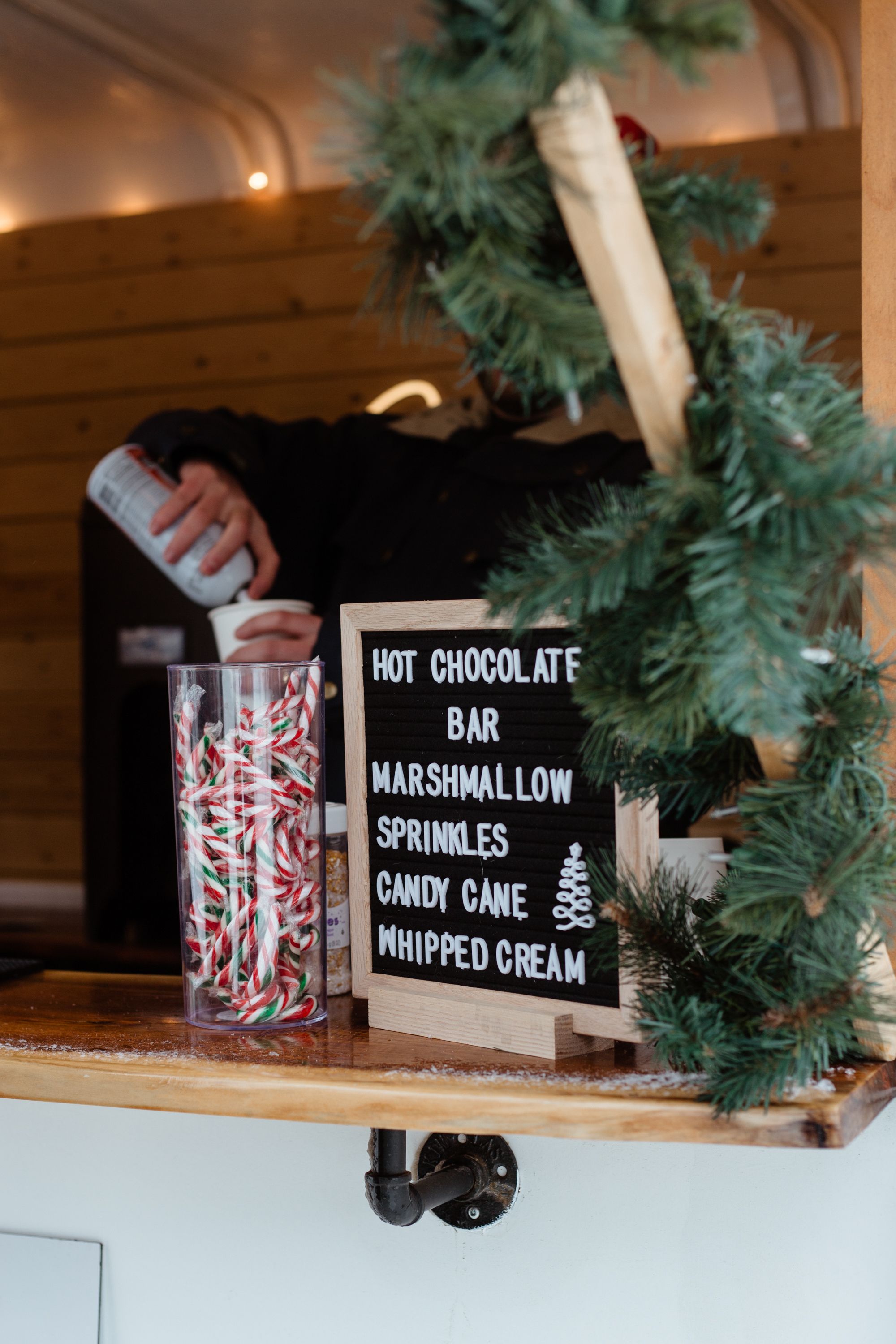 A festive hot chocolate bar with candy canes, marshmallows, and whipped cream toppings displayed on a wooden counter with holiday greenery.
