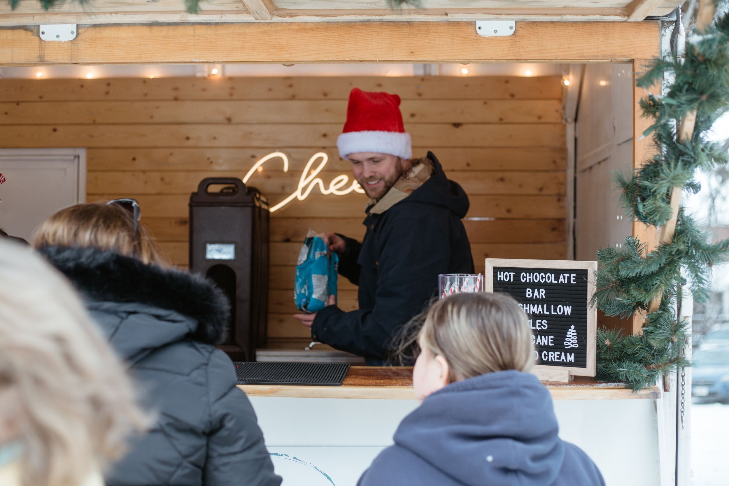 Man in Santa hat serving customers at a hot chocolate bar in a wooden kiosk.