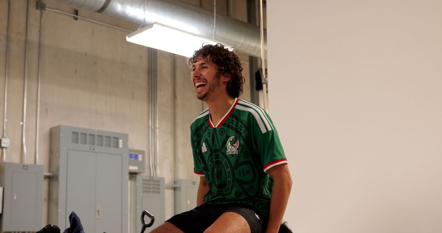 Young man in green Mexico soccer jersey laughing while seated in a bright indoor space.