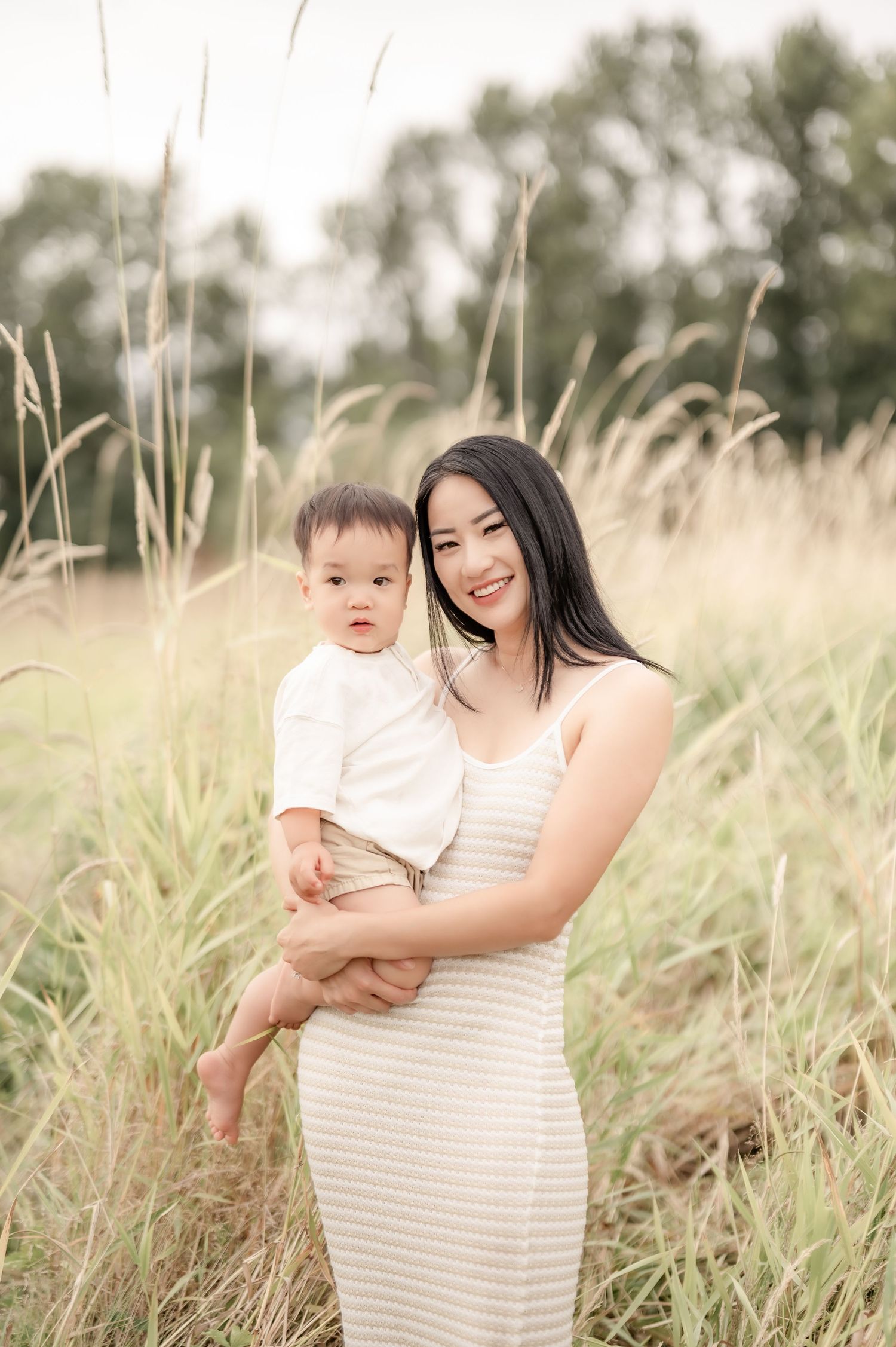 A smiling Asian woman in a striped dress holds a baby boy in a sunlit field of tall golden grass.