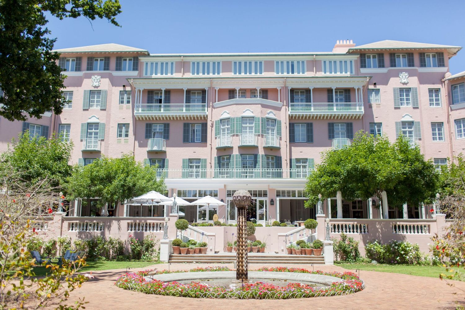 Elegant pink hotel facade with manicured gardens, colorful flower beds, and lush trees on a sunny day.