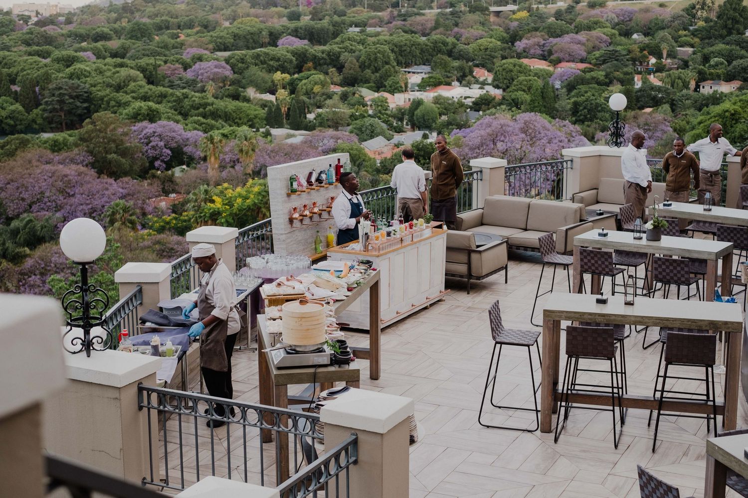 Rooftop terrace bar with white furniture, staff serving guests, and purple jacaranda trees in the background.