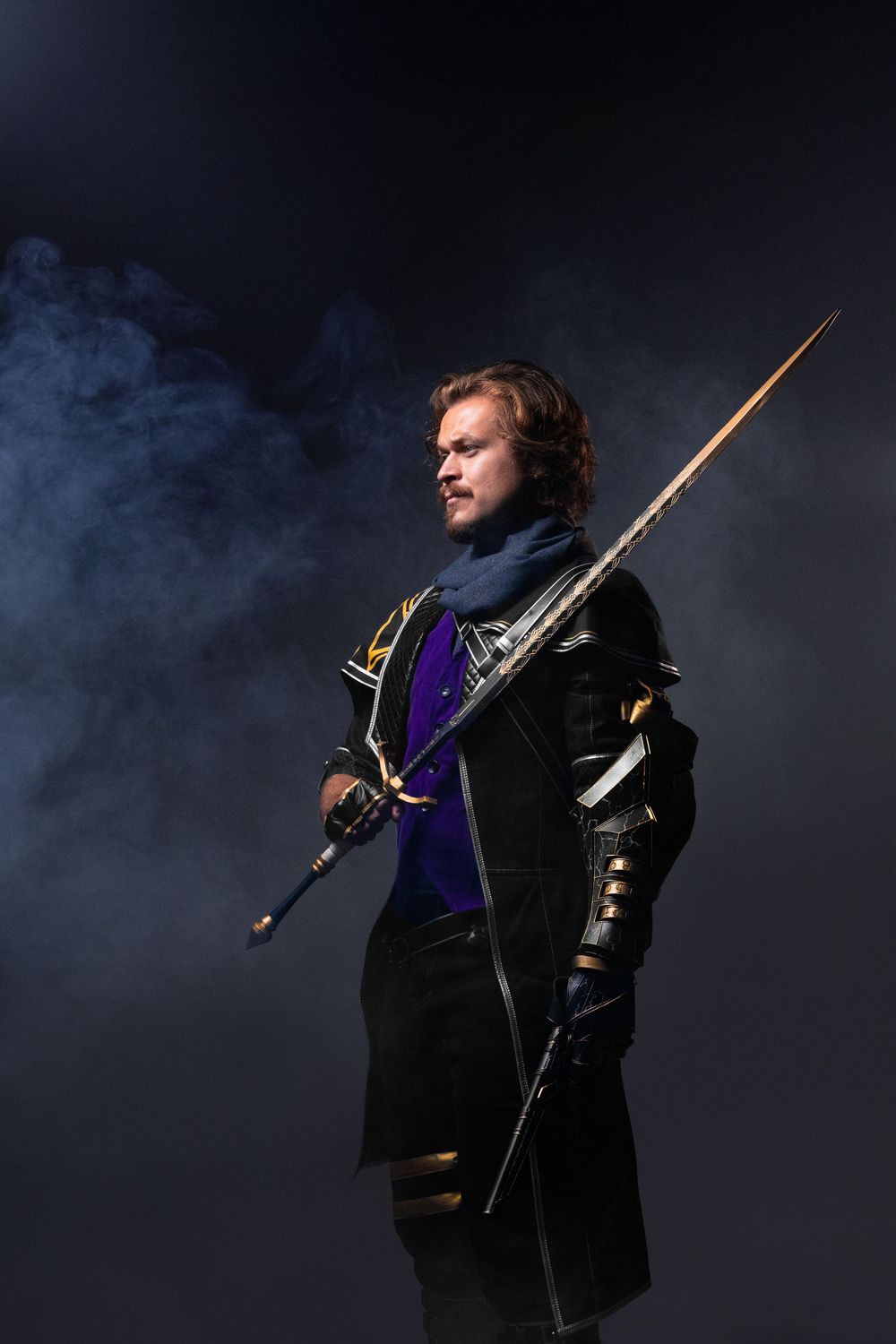 A costumed warrior in dark medieval armor holds a long sword against a smoky black background.