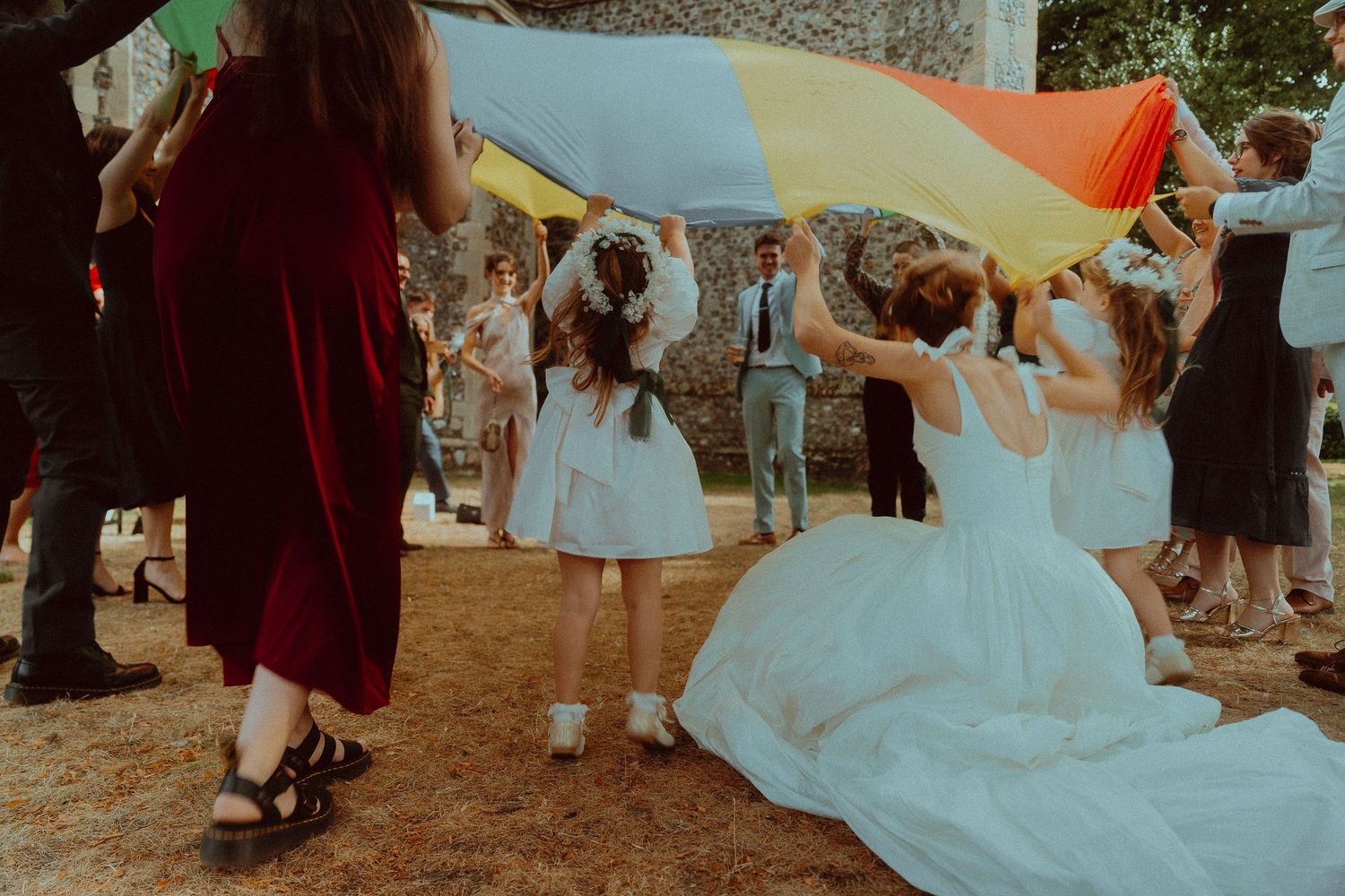 Wedding guests playing with a colorful parachute outdoors, with a bride in a white dress joining the fun.