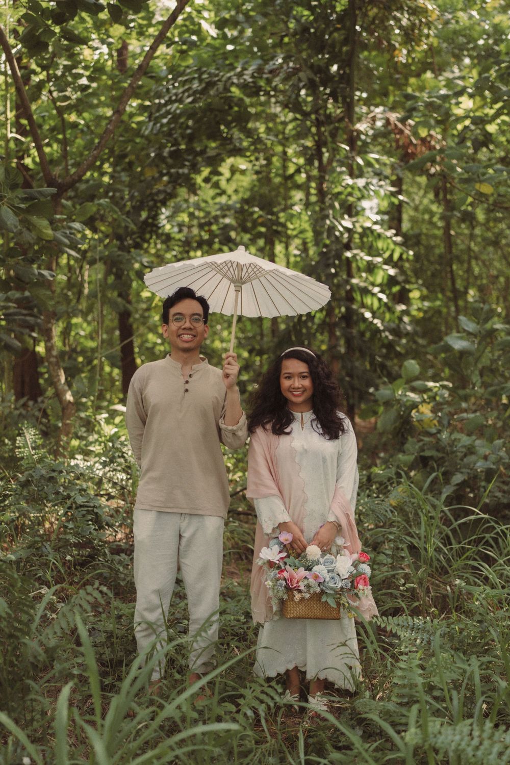 Couple standing in lush green forest, man holding white parasol, woman in pink dress carrying floral bouquet.