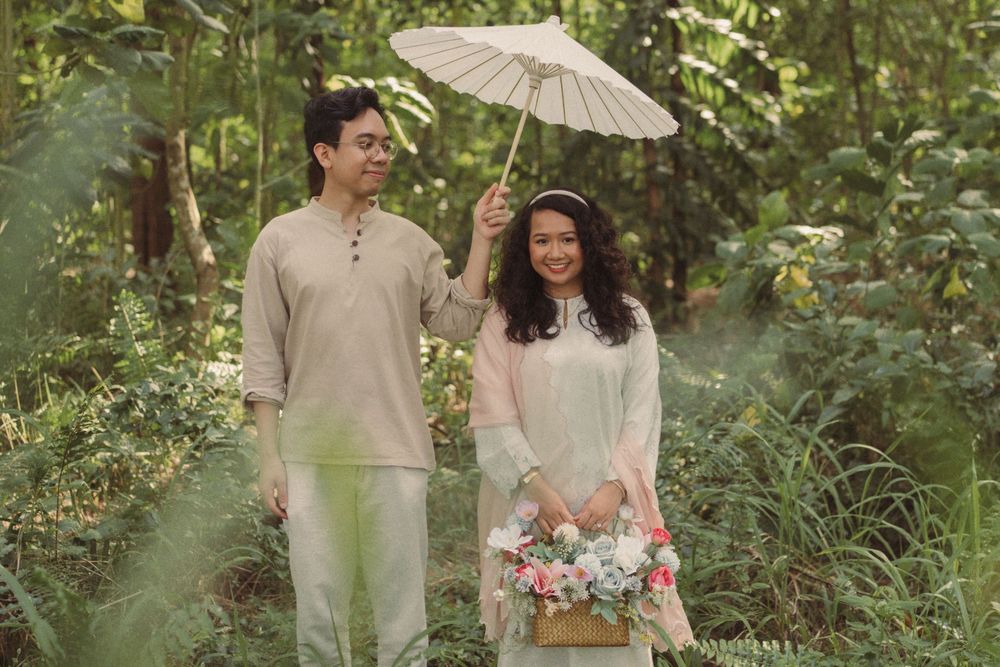 Couple in traditional attire walking through lush forest, man holding white parasol, woman carrying flower basket.