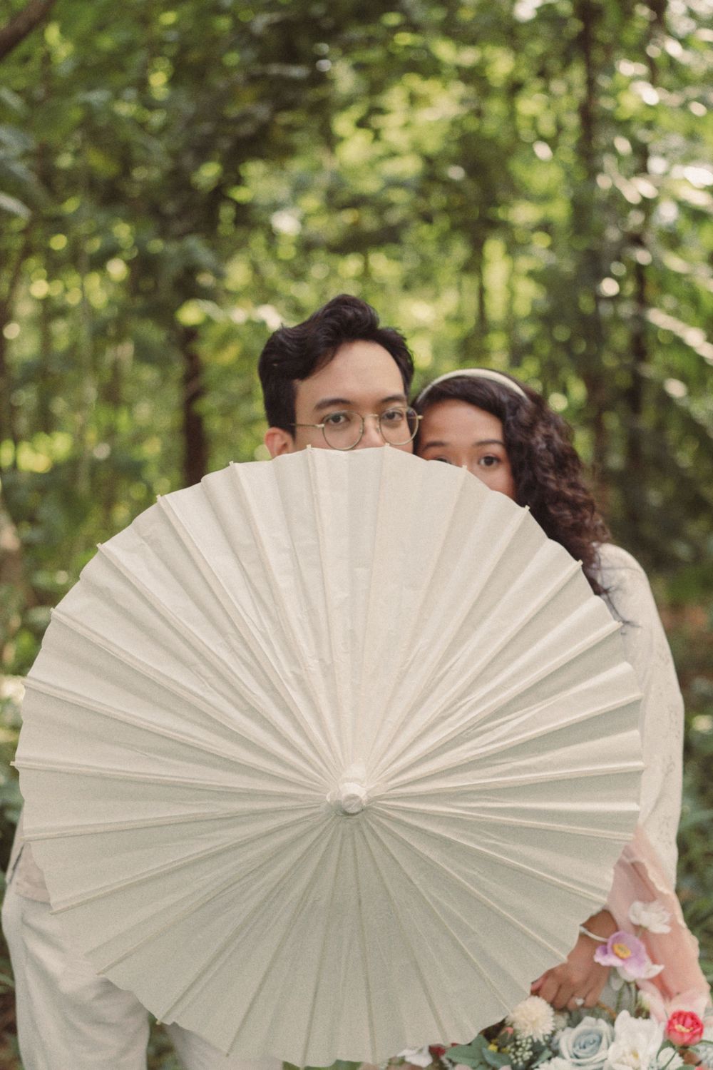 Couple hiding behind a large white paper parasol umbrella in a lush green forest, bride holding a floral bouquet.