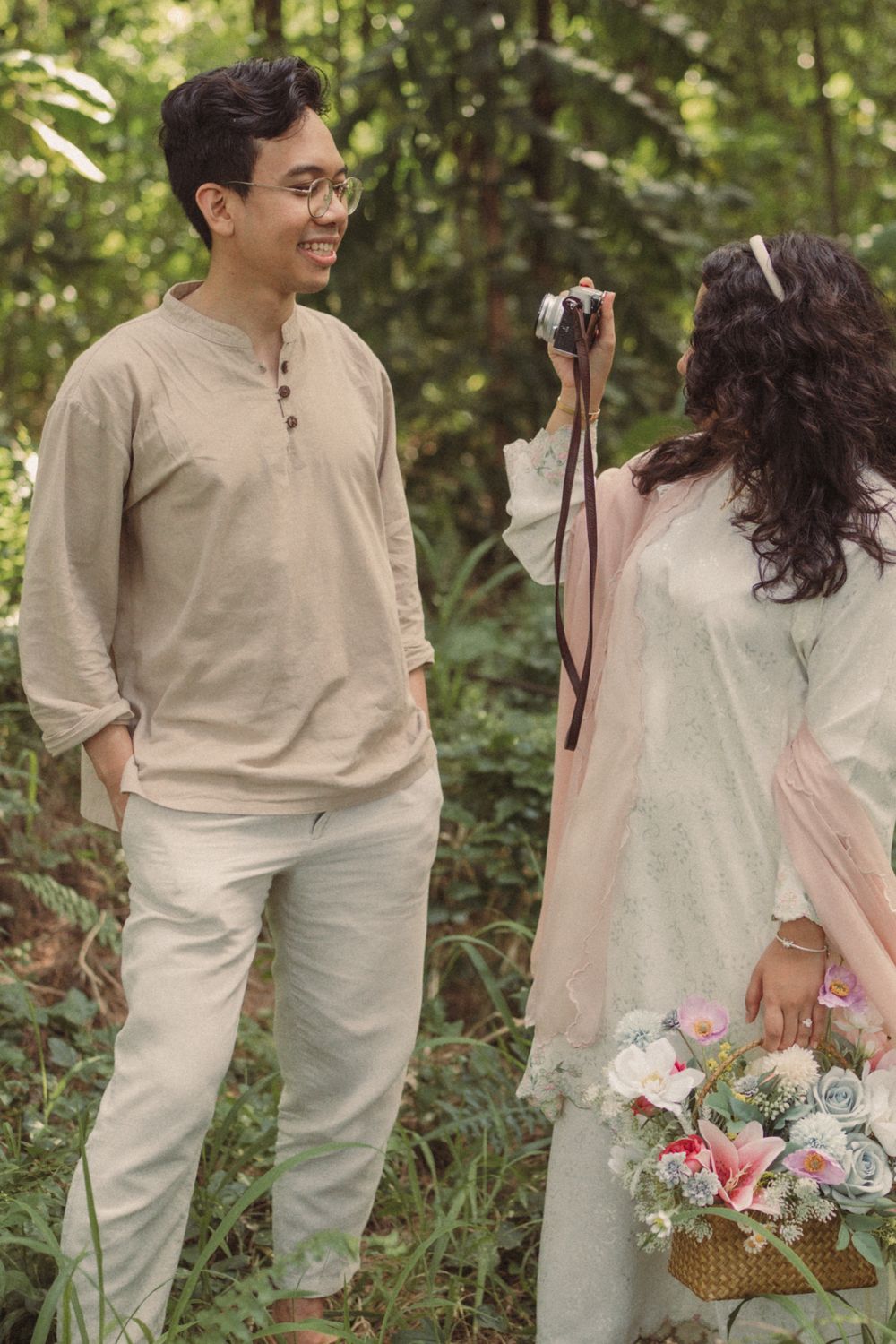 Couple in traditional attire in forest, woman photographing smiling man while holding flower basket.