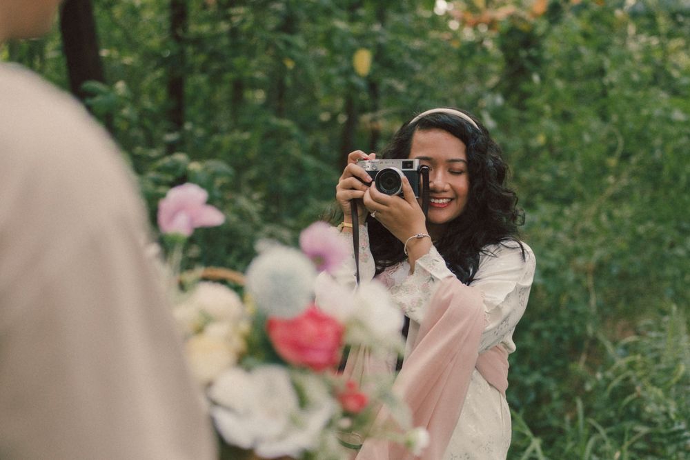 Smiling woman photographing bouquet of flowers with vintage camera outdoors in lush green garden setting.