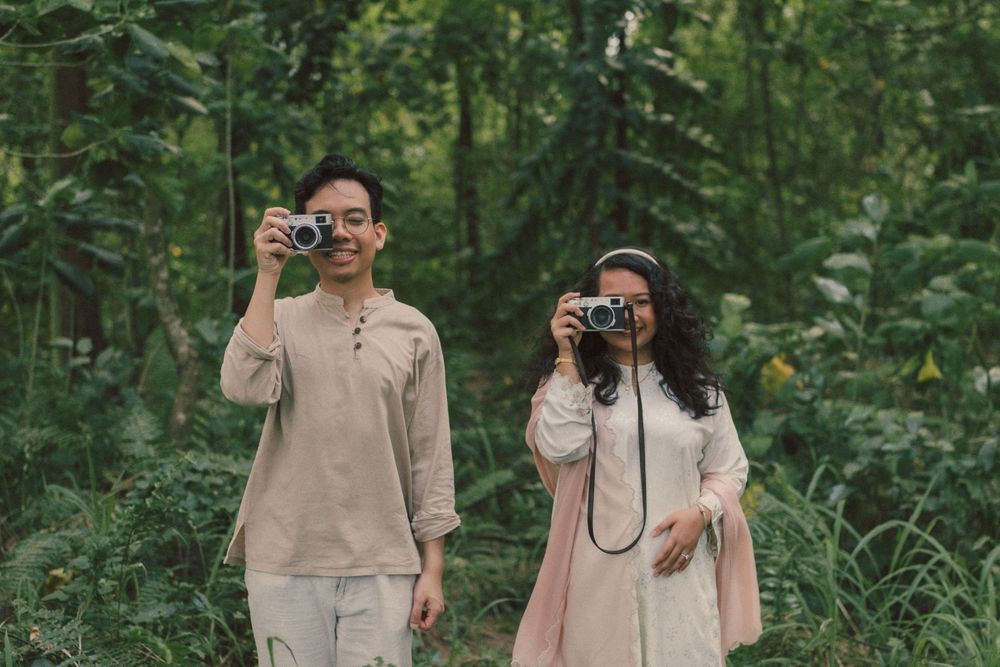 Two people in light clothing hold cameras in a lush green forest, smiling and ready to capture photos.
