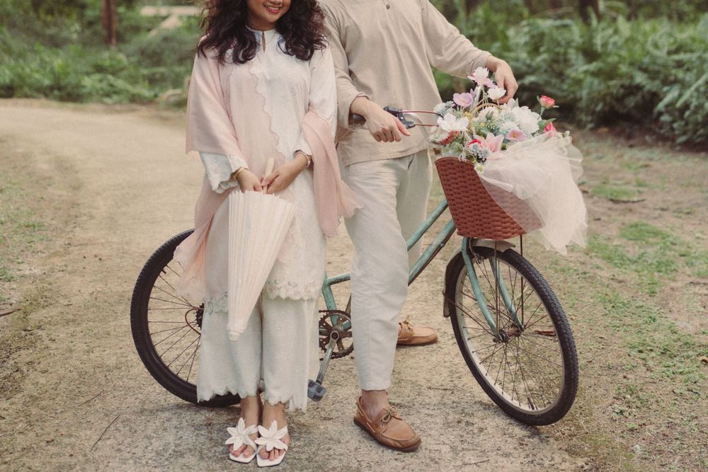 Couple in pastel outfits posing with a vintage bicycle adorned with a flower basket on a serene garden path.