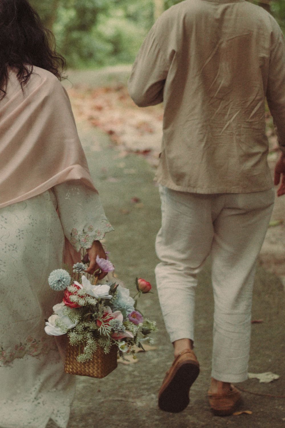 Couple walking outdoors, woman in lace dress holding floral basket bouquet, man in linen outfit, romantic garden stroll.