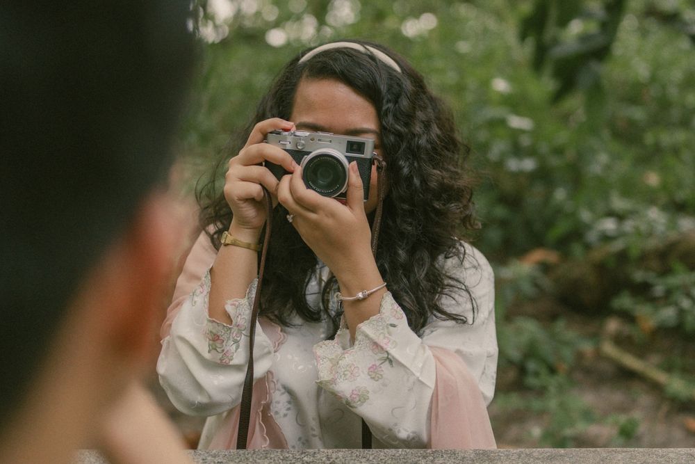 A young woman with curly hair holds a vintage film camera up to her face, photographing outdoors among green foliage.