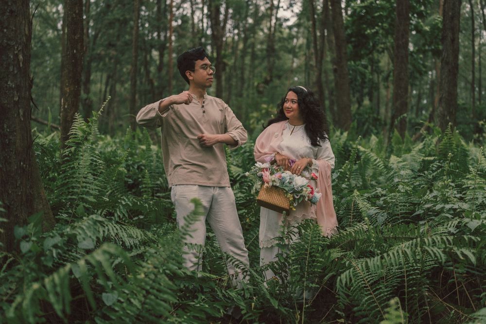 A couple stands among lush green ferns in a forest, the woman holding a floral bouquet in a romantic outdoor setting.