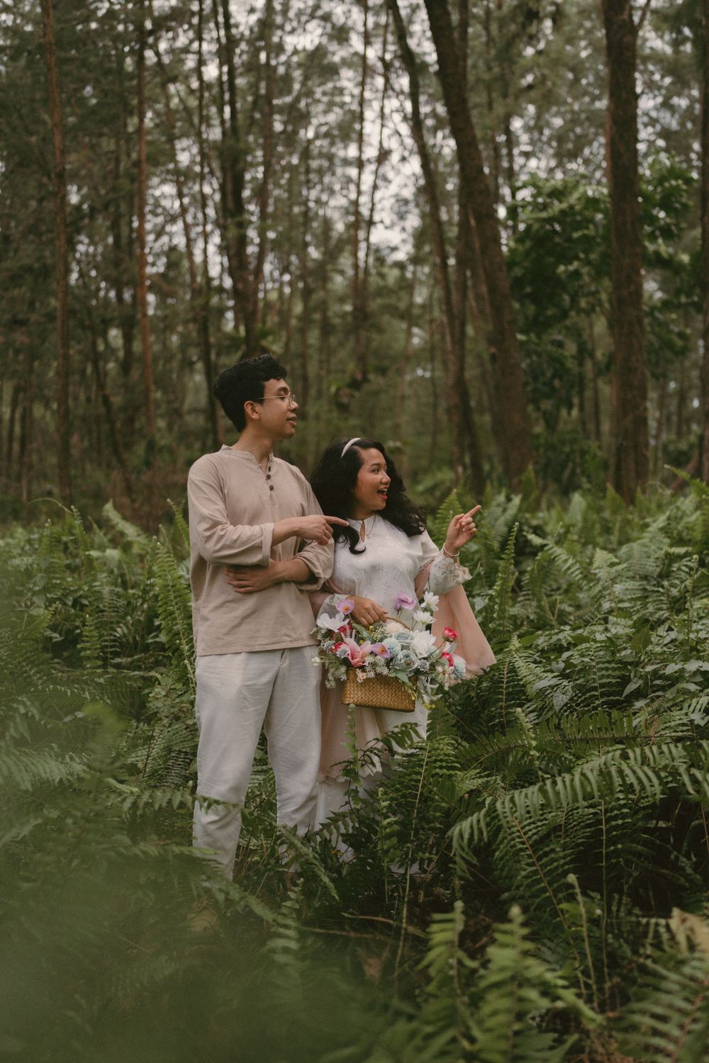 Couple in neutral attire standing among lush ferns in a forest, woman holding a floral bouquet.