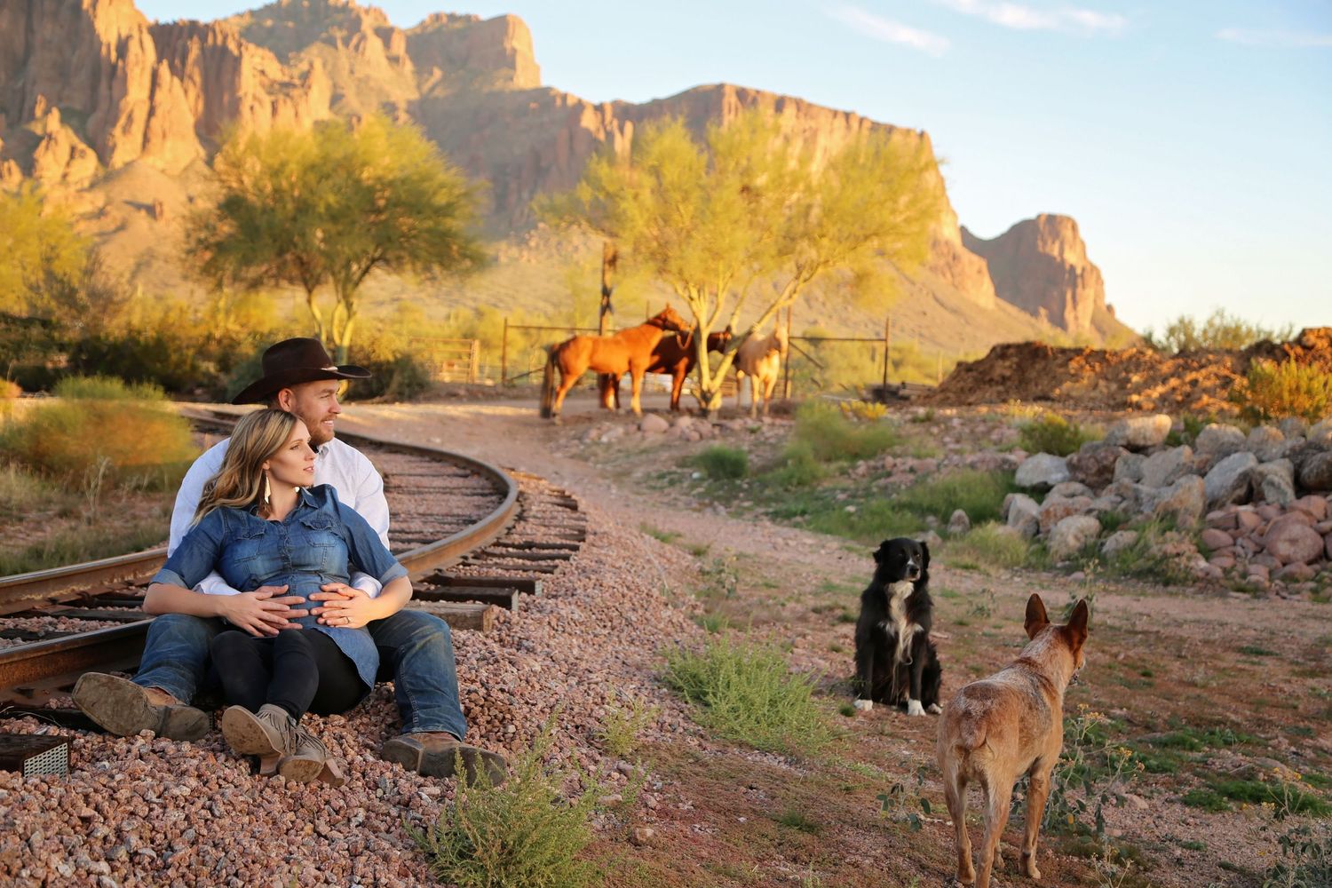 A couple in cowboy hat and denim sit on old railroad tracks with two dogs and horses near desert mountains at golden hour.