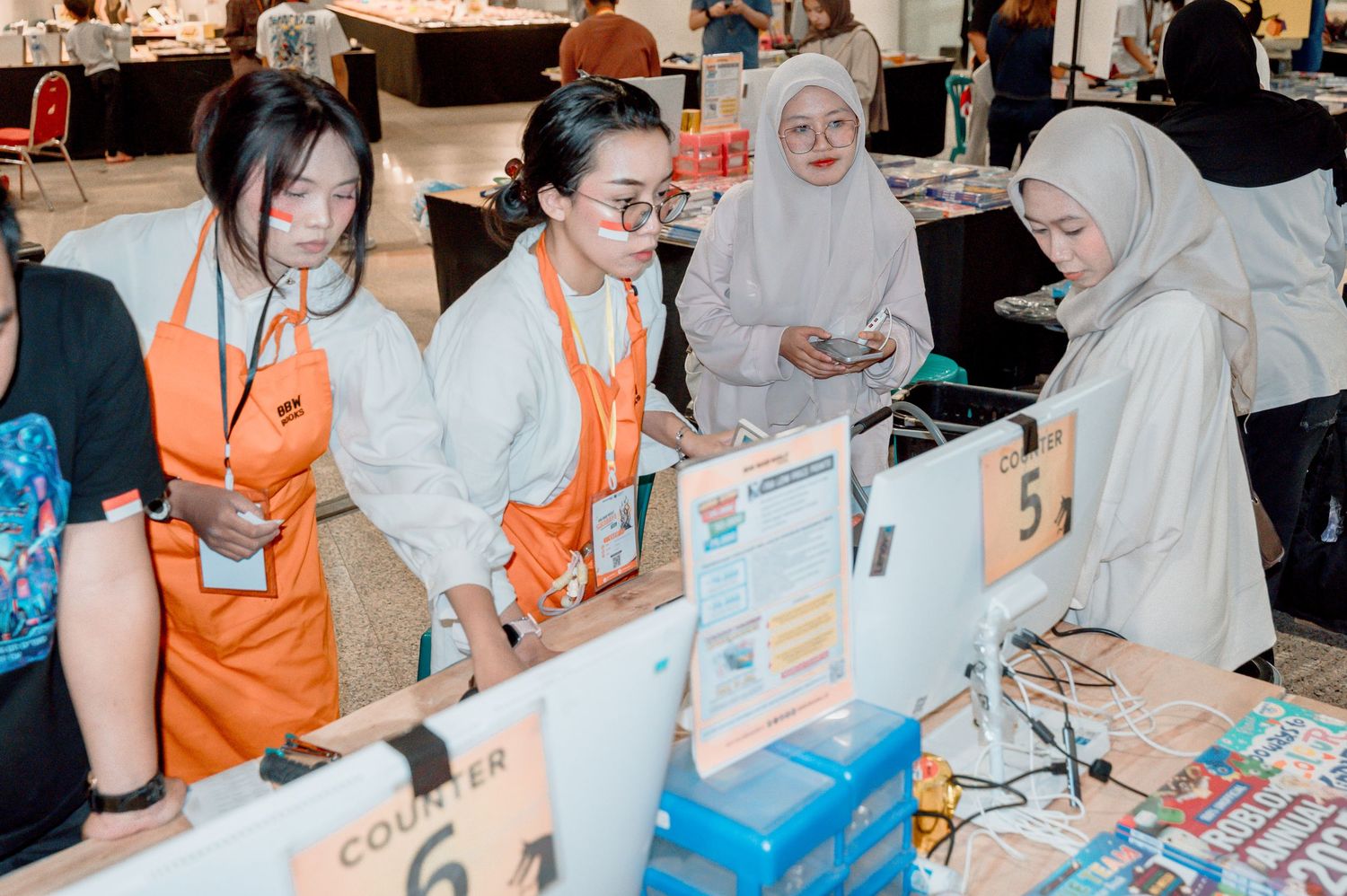 Staff in orange vests assist visitors at a registration counter during an indoor event or exhibition.
