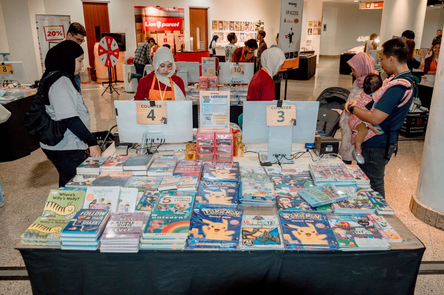 Book fair display tables with Pokemon and educational books, numbered signs 3 and 4, shoppers browsing indoors.