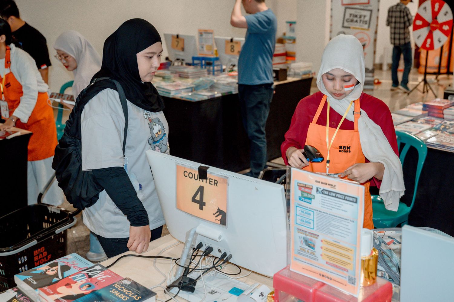 Two women in hijabs at a busy book fair counter, one using a phone, with books and displays visible.