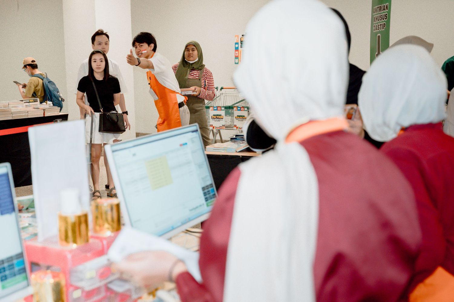 People at an indoor market booth with laptops, perfume bottles, and visitors in hijabs browsing displayed products.