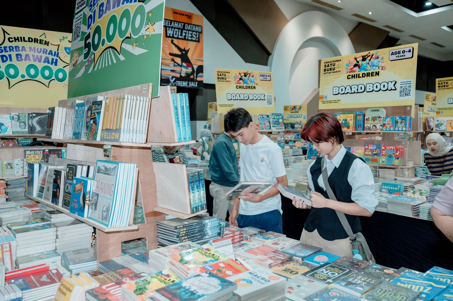 Two shoppers browse colorful children's books and board books at a busy bookstore sale display.