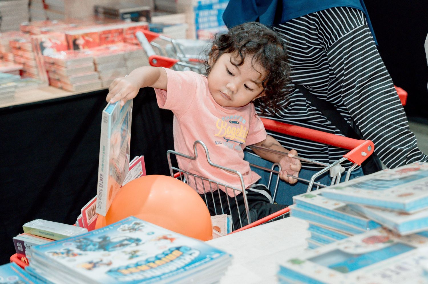 Sleepy toddler in pink shirt sitting in shopping cart reaching for books at a retail store.
