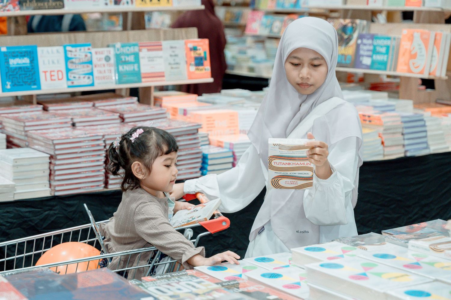 A Muslim mother in hijab shops for books with her young daughter sitting in a shopping cart at a bookstore.